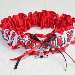 May include: Red and white garter with a black bow and a silver charm in the shape of a hockey stick. The garter has a white background with red lettering that says "Wolverines".