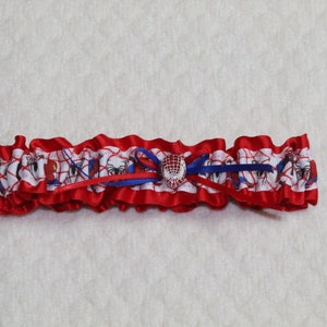 May include: A red and white garter with a blue bow and a spider-man charm. The garter is made of satin and has a ruffled edge.