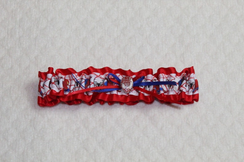 May include: A red and white garter with a blue bow and a spider-man charm. The garter is made of satin and has a ruffled edge.