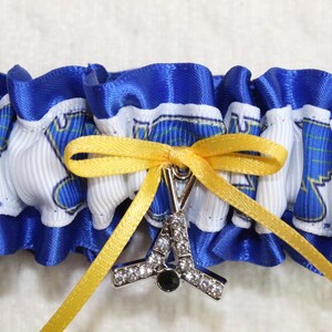 May include: A blue and white ruffled garter featuring a repeating graphic print of a blue winged logo. A yellow ribbon is tied in a bow at the center, holding a silver-toned metal charm shaped like a hockey stick with clear rhinestones.