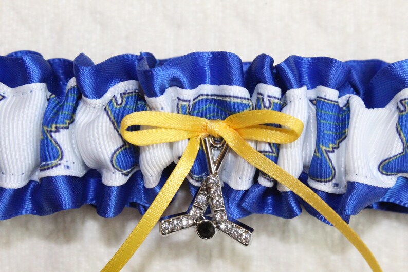 May include: A blue and white ruffled garter featuring a repeating graphic print of a blue winged logo. A yellow ribbon is tied in a bow at the center, holding a silver-toned metal charm shaped like a hockey stick with clear rhinestones.