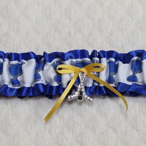 May include: A blue and white ruffled garter featuring a repeating blue musical note pattern on a white ribbon band. The garter is accented with a small yellow satin ribbon bow and a silver-toned charm shaped like a hockey stick with clear crystals.