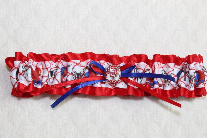 May include: Red and blue garter with a white satin base featuring a repeating pattern of Spiderman characters. A red and blue bow with a Spiderman face button is in the center.