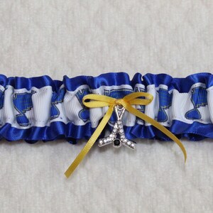 May include: A blue and white ruffled garter featuring a repeating blue musical note pattern on a white ribbon band. The garter is accented with a small yellow satin ribbon bow and a silver-toned charm shaped like a hockey stick with clear crystals.