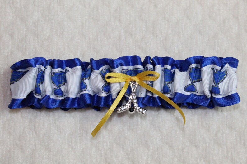 May include: A blue and white ruffled garter featuring a repeating blue musical note pattern on a white ribbon band. The garter is accented with a small yellow satin ribbon bow and a silver-toned charm shaped like a hockey stick with clear crystals.