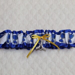 May include: A blue and white ruffled garter featuring a repeating blue logo pattern on a white ribbon base. The garter is accented with a small yellow ribbon bow and a silver-toned charm shaped like a hockey stick encrusted with clear crystals.