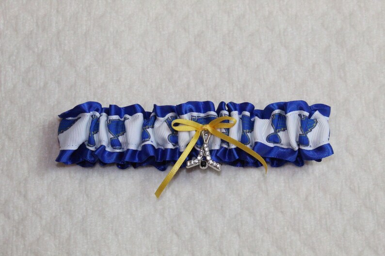 May include: A blue and white ruffled garter featuring a repeating blue logo pattern on a white ribbon base. The garter is accented with a small yellow ribbon bow and a silver-toned charm shaped like a hockey stick encrusted with clear crystals.