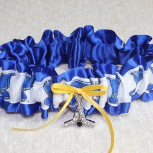 May include: A circular garter featuring a ruffled royal blue satin trim and a white ribbon band with a repeating blue and yellow logo pattern. A small yellow satin bow is centered on the front, accented by a silver-toned charm with clear crystals.