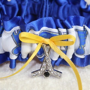 May include: A blue and white ruffled garter featuring a repeating graphic print of a blue lion head. A yellow satin ribbon is tied into a bow at the center, holding a silver-toned charm shaped like two crossed hockey sticks with clear rhinestones.