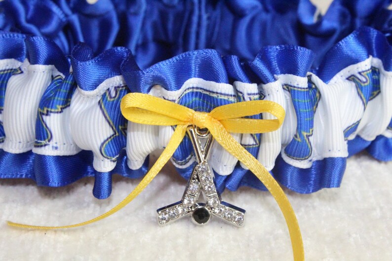 May include: A blue and white ruffled garter featuring a repeating graphic print of a blue lion head. A yellow satin ribbon is tied into a bow at the center, holding a silver-toned charm shaped like two crossed hockey sticks with clear rhinestones.