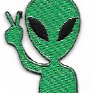 May include: Green alien with black eyes giving the peace sign. The alien is standing with its feet together.