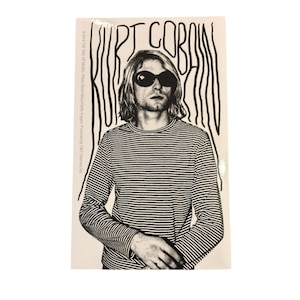 Peut inclure: Autocollant noir et blanc représentant Kurt Cobain portant des lunettes de soleil et un t-shirt à manches longues rayé. Le nom "Kurt Cobain" est écrit au-dessus de l'image dans une police stylisée. L'autocollant est rectangulaire sur fond blanc.
