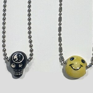 Könnte beinhalten: Zwei Halsketten mit silbernen Kugelkettchen. Eine hat einen schwarzen Totenkopf-Anhänger mit einem weißen Yin-Yang-Symbol. Die andere hat einen gelben Smiley-Anhänger mit schwarzen Augen und einem lächelnden Mund. Beide Anhänger sind aus Keramik.