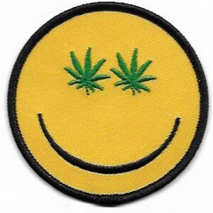 Op de afbeelding: Een gele ronde patch met een zwarte rand. De patch heeft een smiley-gezichtontwerp met groene cannabisbladeren die de ogen vervangen. De mond is een gebogen zwarte lijn.