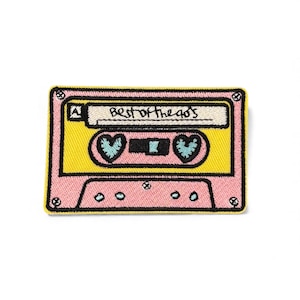Può includere: Toppa rettangolare a forma di musicassetta. La toppa è rosa con il centro giallo. Il testo "BEST OF THE 90'S" è scritto in alto. Due cuori blu sono al centro del nastro.