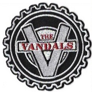 May include: Embroidered patch featuring the text "THE VANDALS" in red, with a large white "V" in the center. The design is set against a black background within a white-edged, gear-shaped border. The patch is likely for clothing or accessories.