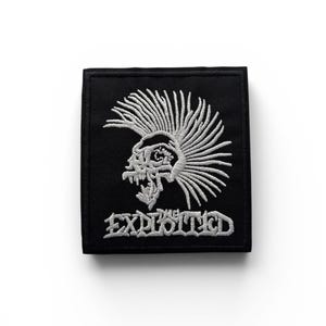 Op de afbeelding: Zwart vierkantige patch met een wit geborduurde schedel met een hanenkam en de tekst "THE EXPLOITED". Deze patch, geschikt voor kleding of accessoires, belichaamt een punkrock-esthetiek.