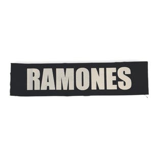 May include: A black rectangular patch featuring the word "RAMONES" in large, white, block lettering. The patch is made of woven fabric, designed to be sewn onto clothing or accessories.