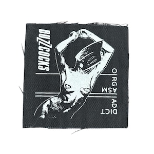 May include: Black square patch with a white illustration of a figure with arms raised and a helmet-like head covering. The text "BUZZCOCKS" is vertically aligned on the left, and "ORG ASM AD DICT" is on the right.