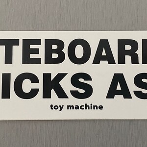 Może przedstawiać: Biała naklejka z czarnym tekstem, kt&oacute;ry brzmi "Skateboarding Kicks Ass" i "toy machine" mniejszą czcionką.
