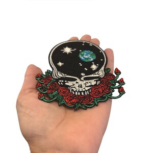 May include: Embroidered patch featuring a skull with a starry night sky design on the head and a floral arrangement of red roses. The patch has a black background and is held in a hand.