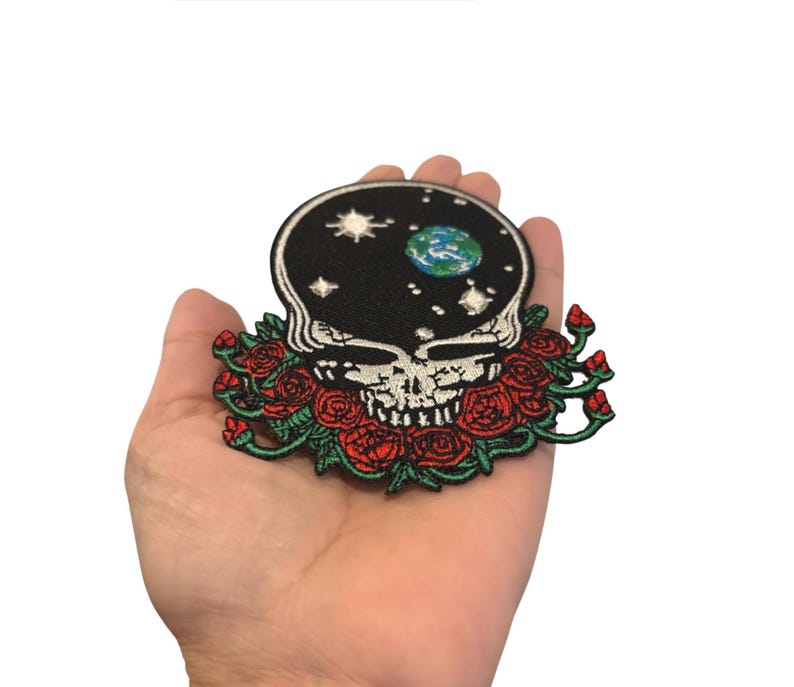 May include: Embroidered patch featuring a skull with a starry night sky design on the head and a floral arrangement of red roses. The patch has a black background and is held in a hand.