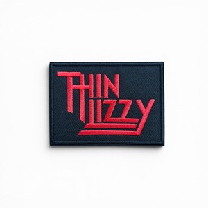 May include: A rectangular embroidered patch with a dark navy blue background and the text Thin Lizzy stitched in bright red thread. The lettering features a stylized, angular font with horizontal lines extending from the base of the letters.