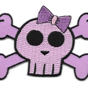 Peut inclure: Un crâne et des os croisés brodés en violet et noir avec un nœud rose sur le dessus.