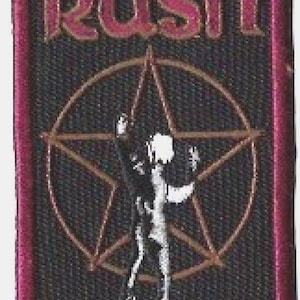 Pode incluir: Patch bordado com o nome da banda "Rush" em letras cor-de-rosa sobre um pentagrama dourado e uma silhueta. O patch tem um fundo preto e uma borda rosa.