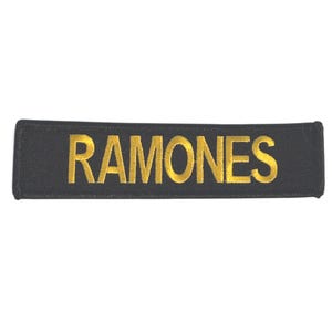 May include: Black rectangular patch with the word "RAMONES" embroidered in gold thread. The patch is a simple, rectangular shape, with the text centered horizontally.