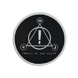 May include: A circular black patch with a white embroidered edge. It displays a white geometric design with an exclamation mark inside a triangle, and the text "PANIC! AT THE DISCO" in white.