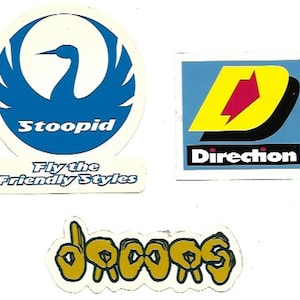 Op de afbeelding: Drie vintage stickers met logo's. De eerste sticker is wit met een blauwe vogel en de tekst "Stoopid Fly the Friendly Styles". De tweede sticker is blauw met een gele "D" en de tekst "Direction". De derde sticker is wit met een geel handgebaar dat "DOOPS" spelt.