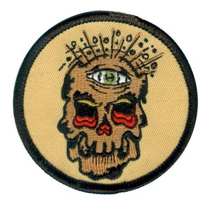 May include: A round embroidered patch with a brown skull with a single green eye and red tear drops. The skull is surrounded by a black circle with a tan background.