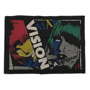 May include: A black embroidered patch with a colourful design. The patch features the words "Vision" and "Aggressor" in white text. The design includes a red, yellow, blue and green abstract face.