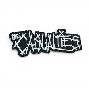 May include: A black patch featuring the white text "THE CASUALTIES". The lettering is in a distressed style. The patch has a jagged border, suitable for application on clothing or accessories.