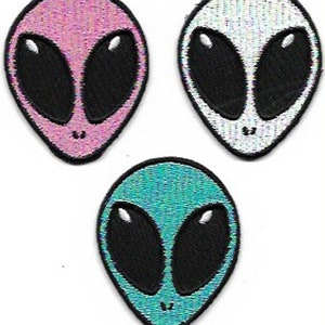May include: Three alien head patches in pink, white, and teal. Each patch features large black oval eyes and a small mouth. The patches are embroidered with a shimmering, iridescent material.