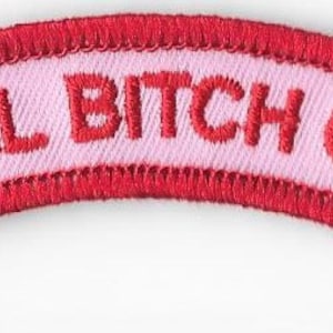 May include: A curved, embroidered patch with the words "TOTAL BITCH CLUB" in red capital letters. The text is on a light pink background, framed by a red border. The patch has a stitched edge.