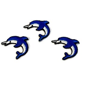 May include: Three blue and white embroidered dolphin patches. The dolphins are facing right and are in a jumping pose.