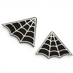 May include: Two black spiderweb-shaped patches with white embroidered detailing. The patches are triangular, with a curved edge and a spiderweb design. The patches are likely for clothing or accessories, and are set against a white background.