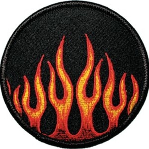 Pode incluir: Um remendo circular preto com um design de chamas vermelhas e amarelas. As chamas são estilizadas e têm uma ponta pontiaguda.