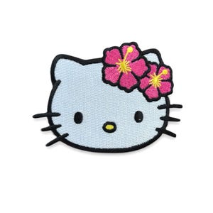 May include: Embroidered patch of a light blue cat face with black whiskers and outline. The cat has a floral crown of pink hibiscus flowers with yellow centres. The patch is on a white background.