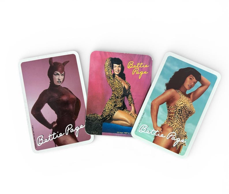 May include: Three vintage-style playing cards featuring images of Bettie Page. The cards showcase pin-up style photos of Bettie Page in various poses and outfits, with the name "Bettie Page" printed on each card.
