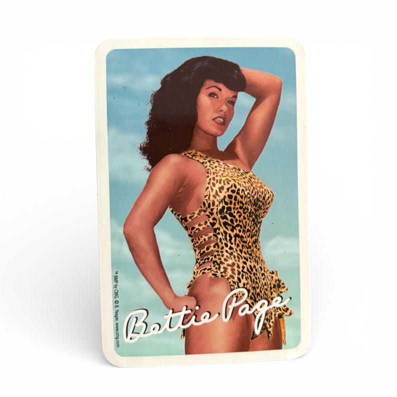 May include: A rectangular card featuring a vintage image of Bettie Page in a leopard print swimsuit. The image has a blue sky background. The text "Bettie Page" is written in a cursive font at the bottom of the card.