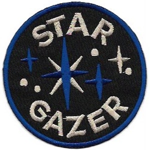 Pode incluir: Patch bordado com as palavras "STAR GAZER" em fio branco sobre fundo preto. O patch apresenta desenhos de estrelas e cruzes azuis, e uma borda azul.