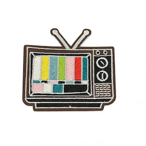 Peut inclure: Écusson brodé représentant un téléviseur vintage. Le téléviseur a un bord marron, un écran affichant des barres verticales colorées et deux antennes argentées. Le côté droit comporte deux boutons et une série de lignes.