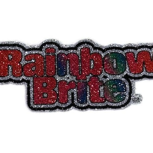 May include: A silver glitter sticker with the words "Rainbow Brite" in red glitter letters. The letters have a rainbow gradient effect.