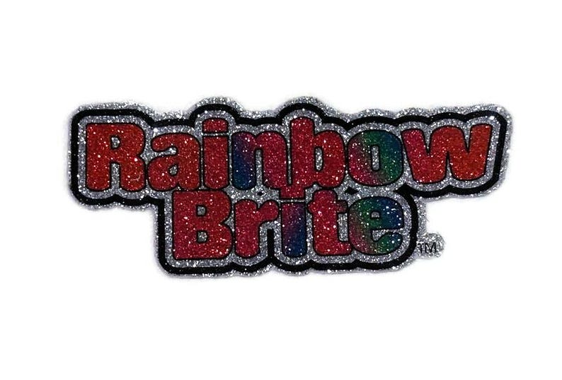 May include: A silver glitter sticker with the words "Rainbow Brite" in red glitter letters. The letters have a rainbow gradient effect.