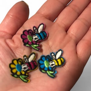 Może przedstawiać: Cztery haftowane naszywki do prasowania z kreskówkowymi pszczołami i kwiatami. Pszczoły są żółte i czarne z białymi skrzydłami. Kwiaty są różowe, niebieskie, żółte i zielone.