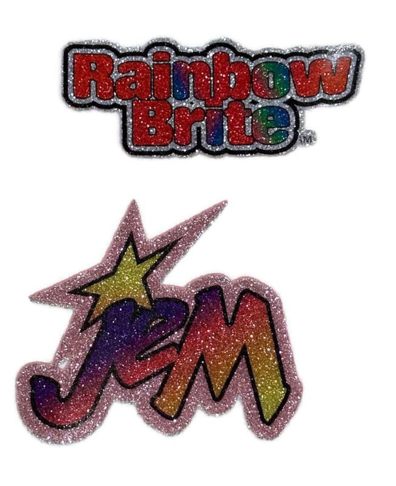 May include: Two iron-on patches with glitter. The first patch is red and silver with the text "Rainbow Brite" in a rainbow font. The second patch is pink and purple with the text "Jem" in a rainbow font with a yellow star.