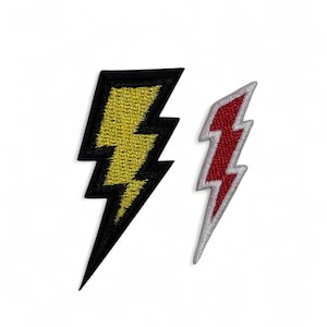 Könnte beinhalten: Zwei gestickte Aufnäher in Form von Blitzen. Einer ist schwarz mit gelber Mitte, der andere ist rot mit silbernem Rand. Die Aufnäher sind wahrscheinlich für Kleidung oder Accessoires.